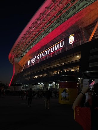 An illuminated stadium with a large neon sign reading 'nef Stadyumu' on the exterior of the building. The stadium is well-lit with red and white lights, creating a vibrant atmosphere. The structure is modern with large glass windows and a wide entrance area. A crowd of people is gathered outside, and a person in the foreground is taking a photo with their smartphone.