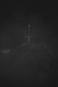 A sword is embedded in a large stone or rock, shrouded in darkness and mist. The overall image is dimly lit, creating a mysterious and dramatic atmosphere.
