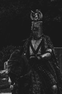 A medieval knight wearing a detailed suit of armor, including a helmet with a mermaid figure on top, sits atop a horse. The horse is also covered in protective gear. The image is in black and white, emphasizing the textures and patterns of the metal and cloth.