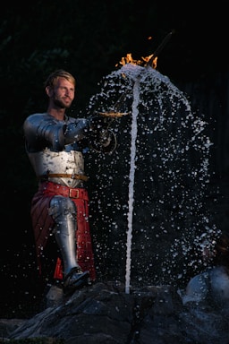 A person wearing medieval full-body armor stands holding a sword. Water splashes dramatically around the blade, and the top of the sword is engulfed in flames. The background is dark, highlighting the figure and the elements in motion.