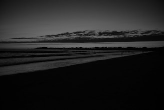 A monochrome image captures a serene beach at dusk, with a vast expanse of dark water meeting the shoreline. The sky is overcast with a layer of clouds, with a faint glow on the horizon indicating either sunrise or sunset. Silhouettes of distant people are visible along the water’s edge.