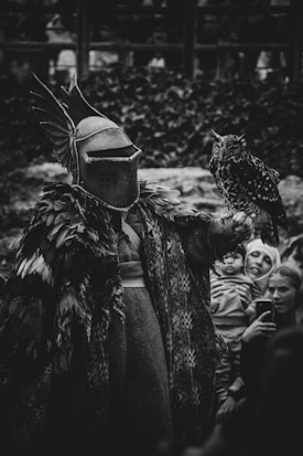 A medieval knight in elaborate armor adorned with feathers holds an owl. The background is filled with a crowd of onlookers, capturing the scene in black and white.