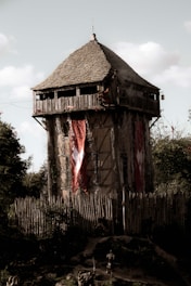 A medieval-style wooden watchtower with a conical roof stands tall, surrounded by a palisade fence made of wooden stakes. Large red banners with white emblems hang from the sides of the tower. The scene has a medieval or historical atmosphere, with a few figures on the ground dressed in dark, armor-like clothing. The tower is set against a backdrop of trees and a partly cloudy sky.