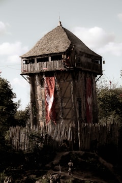 A medieval-style wooden watchtower with a conical roof stands tall, surrounded by a palisade fence made of wooden stakes. Large red banners with white emblems hang from the sides of the tower. The scene has a medieval or historical atmosphere, with a few figures on the ground dressed in dark, armor-like clothing. The tower is set against a backdrop of trees and a partly cloudy sky.