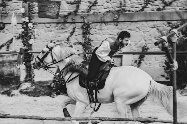 A person in medieval attire rides a galloping white horse in an outdoor setting, surrounded by stone walls and rustic elements. The scene captures a sense of movement and action, with the rider appearing focused and intense.
