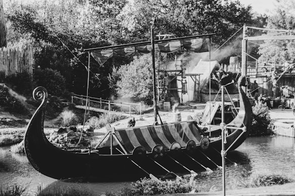 A detailed black-and-white image shows a large Viking ship docked on a tranquil body of water. The ship features a distinctive curved bow and stern, adorned with shields and oars. Surrounding the vessel, lush greenery and rustic wooden fencing add to the historical setting. A partially hidden structure resembling a thatched-roof building is visible in the background, suggesting a traditional village or settlement scene.