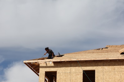 A person is working on the roof of a wooden building under construction, using a tool. The sky is bright and partly cloudy, creating a strong contrast with the wooden structure.