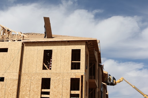 A multi-story wooden building under construction, with a worker on the roof holding a large wooden board. A crane is positioned on the side, assisting with the construction. The sky is bright and partly cloudy.