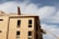 A multi-story wooden building under construction, with a worker on the roof holding a large wooden board. A crane is positioned on the side, assisting with the construction. The sky is bright and partly cloudy.