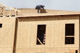 A construction worker is on the roof of a partially built wooden house, handling a piece of plywood. The wooden structure is in the initial stages of development and consists mainly of a framed outer wall without siding or windows. There is a ladder inside the building, visible through an opening.