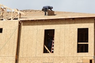 A construction worker is on the roof of a partially built wooden house, handling a piece of plywood. The wooden structure is in the initial stages of development and consists mainly of a framed outer wall without siding or windows. There is a ladder inside the building, visible through an opening.