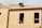 A construction worker is on the roof of a partially built wooden house, handling a piece of plywood. The wooden structure is in the initial stages of development and consists mainly of a framed outer wall without siding or windows. There is a ladder inside the building, visible through an opening.
