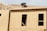 A construction worker is on the roof of a partially built wooden house, handling a piece of plywood. The wooden structure is in the initial stages of development and consists mainly of a framed outer wall without siding or windows. There is a ladder inside the building, visible through an opening.