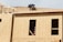 A construction worker is on the roof of a partially built wooden house, handling a piece of plywood. The wooden structure is in the initial stages of development and consists mainly of a framed outer wall without siding or windows. There is a ladder inside the building, visible through an opening.
