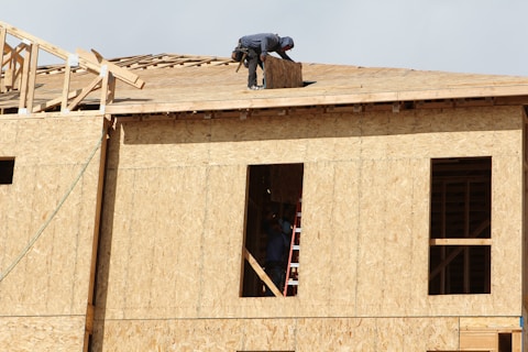 A construction worker is on the roof of a partially built wooden house, handling a piece of plywood. The wooden structure is in the initial stages of development and consists mainly of a framed outer wall without siding or windows. There is a ladder inside the building, visible through an opening.