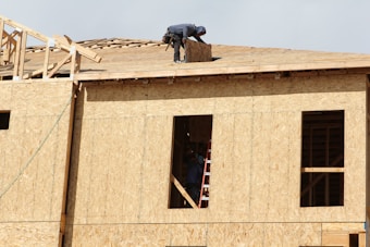 A construction worker is on the roof of a partially built wooden house, handling a piece of plywood. The wooden structure is in the initial stages of development and consists mainly of a framed outer wall without siding or windows. There is a ladder inside the building, visible through an opening.