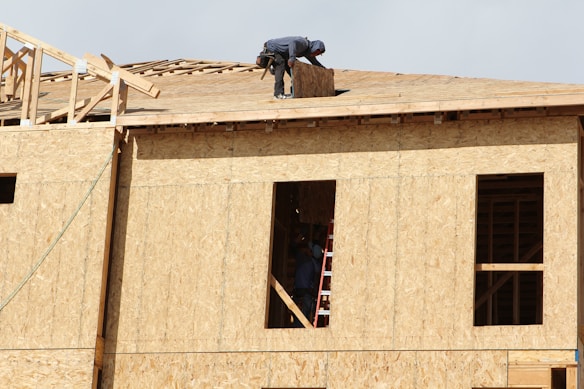 A construction worker is on the roof of a partially built wooden house, handling a piece of plywood. The wooden structure is in the initial stages of development and consists mainly of a framed outer wall without siding or windows. There is a ladder inside the building, visible through an opening.