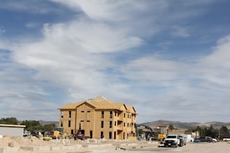 A multi-story building under construction with wooden framing is situated in an open area. Various construction vehicles, including excavators and trucks, are present around the site. The sky is partly cloudy, and there are hills or mountains in the distant background.