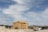 A multi-story building under construction with wooden framing is situated in an open area. Various construction vehicles, including excavators and trucks, are present around the site. The sky is partly cloudy, and there are hills or mountains in the distant background.