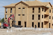 A construction site with a multi-story wooden building frame in progress. There's an excavator with a red arm and a person operating it near a pile of construction materials. Another worker is also visible, wearing a hard hat and is engaged in groundwork.