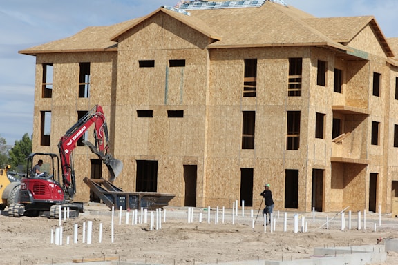 A construction site with a multi-story wooden building frame in progress. There's an excavator with a red arm and a person operating it near a pile of construction materials. Another worker is also visible, wearing a hard hat and is engaged in groundwork.