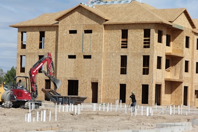 A construction site with a multi-story wooden building frame in progress. There's an excavator with a red arm and a person operating it near a pile of construction materials. Another worker is also visible, wearing a hard hat and is engaged in groundwork.
