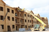 A partially constructed wooden building is visible, where workers are using a yellow telescopic forklift to lift materials to the roof. The building has multiple floors with evenly spaced window openings, and several wooden beams and construction materials are scattered around the site. The sky appears clear with some clouds, and the overall scene suggests an active construction site.