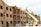 A partially constructed wooden building is visible, where workers are using a yellow telescopic forklift to lift materials to the roof. The building has multiple floors with evenly spaced window openings, and several wooden beams and construction materials are scattered around the site. The sky appears clear with some clouds, and the overall scene suggests an active construction site.