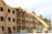 A partially constructed wooden building is visible, where workers are using a yellow telescopic forklift to lift materials to the roof. The building has multiple floors with evenly spaced window openings, and several wooden beams and construction materials are scattered around the site. The sky appears clear with some clouds, and the overall scene suggests an active construction site.