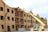 A partially constructed wooden building is visible, where workers are using a yellow telescopic forklift to lift materials to the roof. The building has multiple floors with evenly spaced window openings, and several wooden beams and construction materials are scattered around the site. The sky appears clear with some clouds, and the overall scene suggests an active construction site.