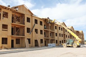 A multi-story wooden building under construction with an open framework. A yellow construction vehicle with a crane attachment lifts workers towards the upper levels. The building features multiple window openings, and construction materials are scattered around the site. The sky is partly cloudy, adding to the scene of active construction work.