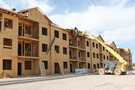 A multi-story wooden building under construction with an open framework. A yellow construction vehicle with a crane attachment lifts workers towards the upper levels. The building features multiple window openings, and construction materials are scattered around the site. The sky is partly cloudy, adding to the scene of active construction work.