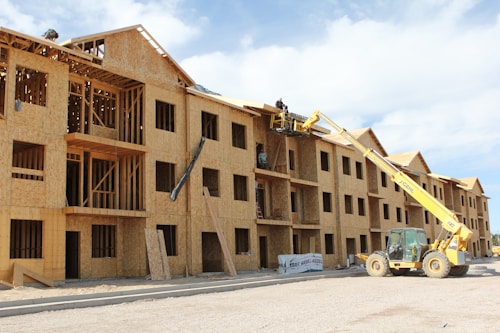 A multi-story wooden building under construction with an open framework. A yellow construction vehicle with a crane attachment lifts workers towards the upper levels. The building features multiple window openings, and construction materials are scattered around the site. The sky is partly cloudy, adding to the scene of active construction work.