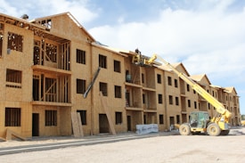 A multi-story wooden building under construction with an open framework. A yellow construction vehicle with a crane attachment lifts workers towards the upper levels. The building features multiple window openings, and construction materials are scattered around the site. The sky is partly cloudy, adding to the scene of active construction work.