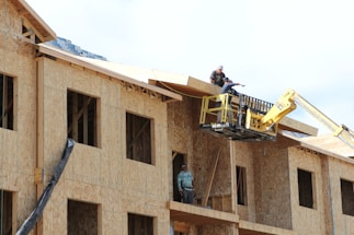 A construction scene with workers building a multi-story wooden structure. Two people are on the roof and another is on a lift, all actively engaged in the building process. The unfinished structure has exposed wooden frames and large window openings.