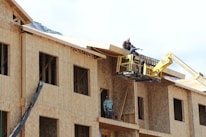 A construction scene with workers building a multi-story wooden structure. Two people are on the roof and another is on a lift, all actively engaged in the building process. The unfinished structure has exposed wooden frames and large window openings.