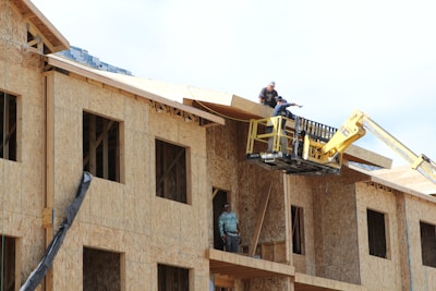 A construction scene with workers building a multi-story wooden structure. Two people are on the roof and another is on a lift, all actively engaged in the building process. The unfinished structure has exposed wooden frames and large window openings.