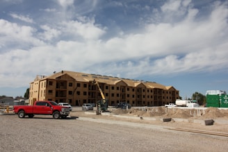 A large, multi-story building under construction with wooden framing visible. Several construction vehicles, including a red truck and a crane, are parked nearby. Piles of dirt and construction materials are scattered around the site under a partly cloudy sky.