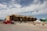 A large, multi-story building under construction with wooden framing visible. Several construction vehicles, including a red truck and a crane, are parked nearby. Piles of dirt and construction materials are scattered around the site under a partly cloudy sky.