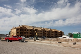 A large, multi-story building under construction with wooden framing visible. Several construction vehicles, including a red truck and a crane, are parked nearby. Piles of dirt and construction materials are scattered around the site under a partly cloudy sky.