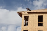 A partially constructed building made of plywood panels is being worked on by a person kneeling on the roof. Another person is visible through the window frames, presumably assisting with the construction. The sky is largely clear with some clouds.