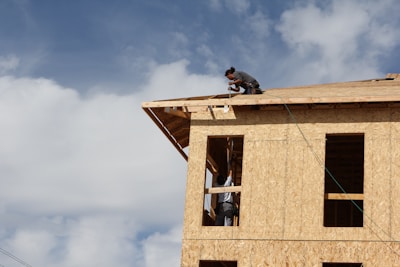 A partially constructed building made of plywood panels is being worked on by a person kneeling on the roof. Another person is visible through the window frames, presumably assisting with the construction. The sky is largely clear with some clouds.