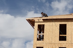 A partially constructed building made of plywood panels is being worked on by a person kneeling on the roof. Another person is visible through the window frames, presumably assisting with the construction. The sky is largely clear with some clouds.