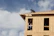 A partially constructed building made of plywood panels is being worked on by a person kneeling on the roof. Another person is visible through the window frames, presumably assisting with the construction. The sky is largely clear with some clouds.