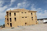 A large multi-story building under construction with wooden framework and sheathing. Workers can be seen on the roof. The surrounding area features construction materials and vehicles. The sky is clear with a few clouds.