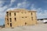 A large multi-story building under construction with wooden framework and sheathing. Workers can be seen on the roof. The surrounding area features construction materials and vehicles. The sky is clear with a few clouds.