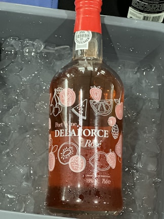 A bottle of Port Wine labeled 'DELAFORCE' with 'Rosé' below it is placed in a container filled with ice. The bottle has a red cap and is adorned with illustrations of various fruits such as cherries, watermelon slices, strawberries, oranges, and kiwis. There are visible liquid droplets on the bottle due to the cold ice.