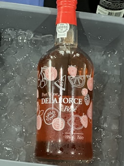 A bottle of Port Wine labeled 'DELAFORCE' with 'Rosé' below it is placed in a container filled with ice. The bottle has a red cap and is adorned with illustrations of various fruits such as cherries, watermelon slices, strawberries, oranges, and kiwis. There are visible liquid droplets on the bottle due to the cold ice.