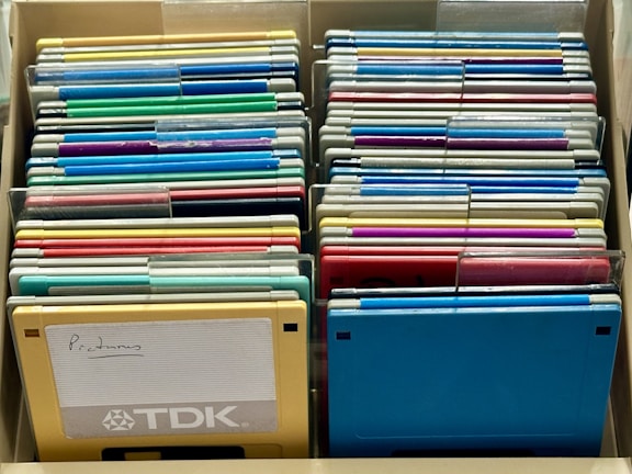 A collection of colorful floppy disks is organized in a storage box. The disks come in various colors, including blue, green, yellow, and red. A prominent yellow disk in the front has the word 'Programs' handwritten on a label and features the TDK branding.