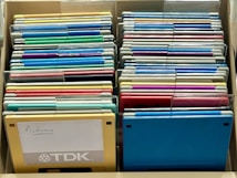 A collection of colorful floppy disks is organized in a storage box. The disks come in various colors, including blue, green, yellow, and red. A prominent yellow disk in the front has the word 'Programs' handwritten on a label and features the TDK branding.